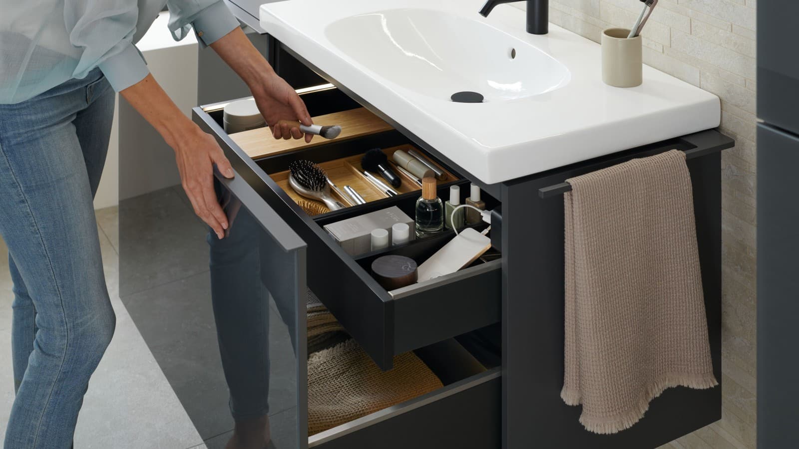 Woman reaches for a brush from an open Geberit Acanto washbasin cabinet in a lava finish Woman reaches for a brush from an open Geberit Acanto washbasin cabinet in a lava finish